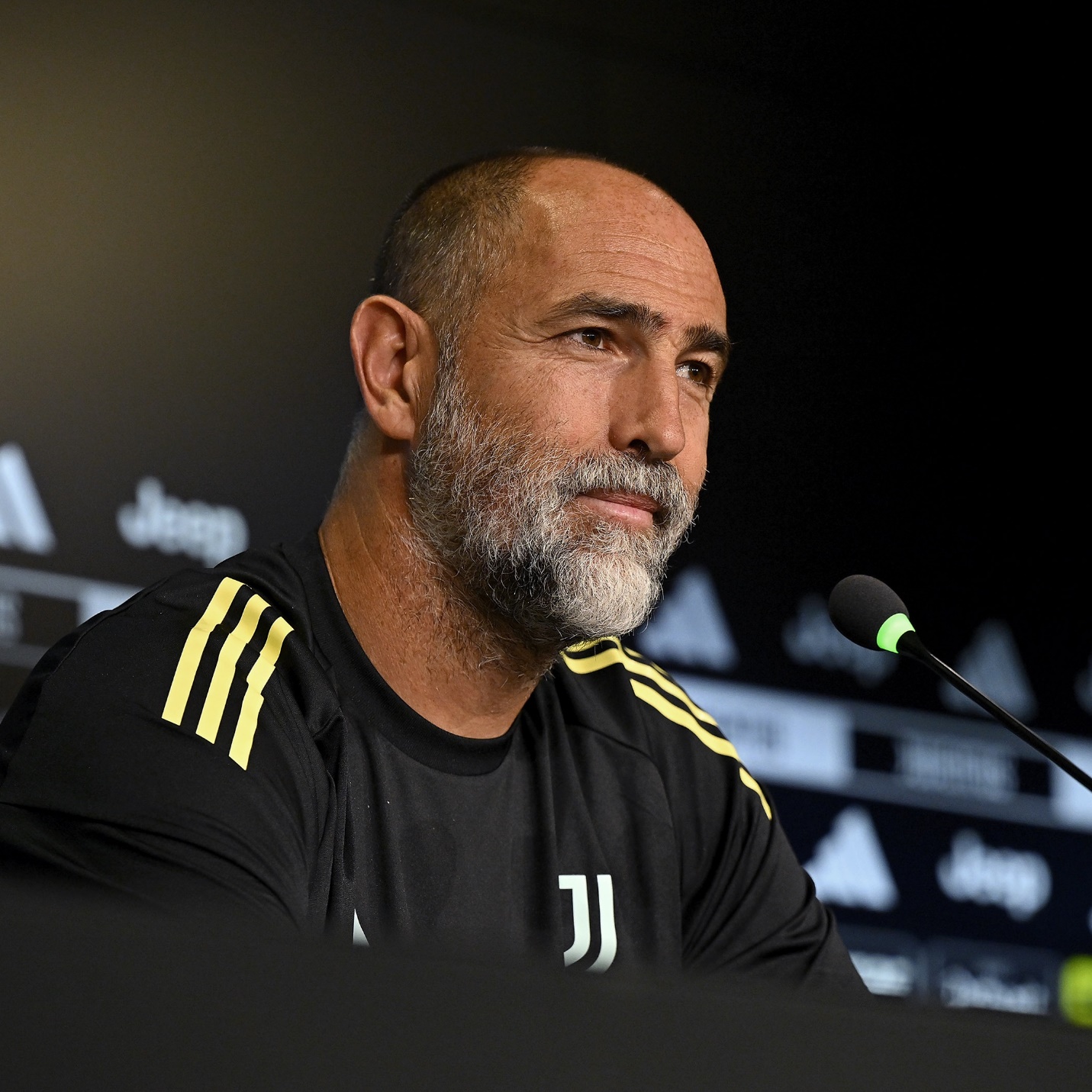 A man with a bald head and gray beard wearing a black suit white shirt and black tie stands with arms crossed in front of a blurred stadium crowd background with red and white flags visible